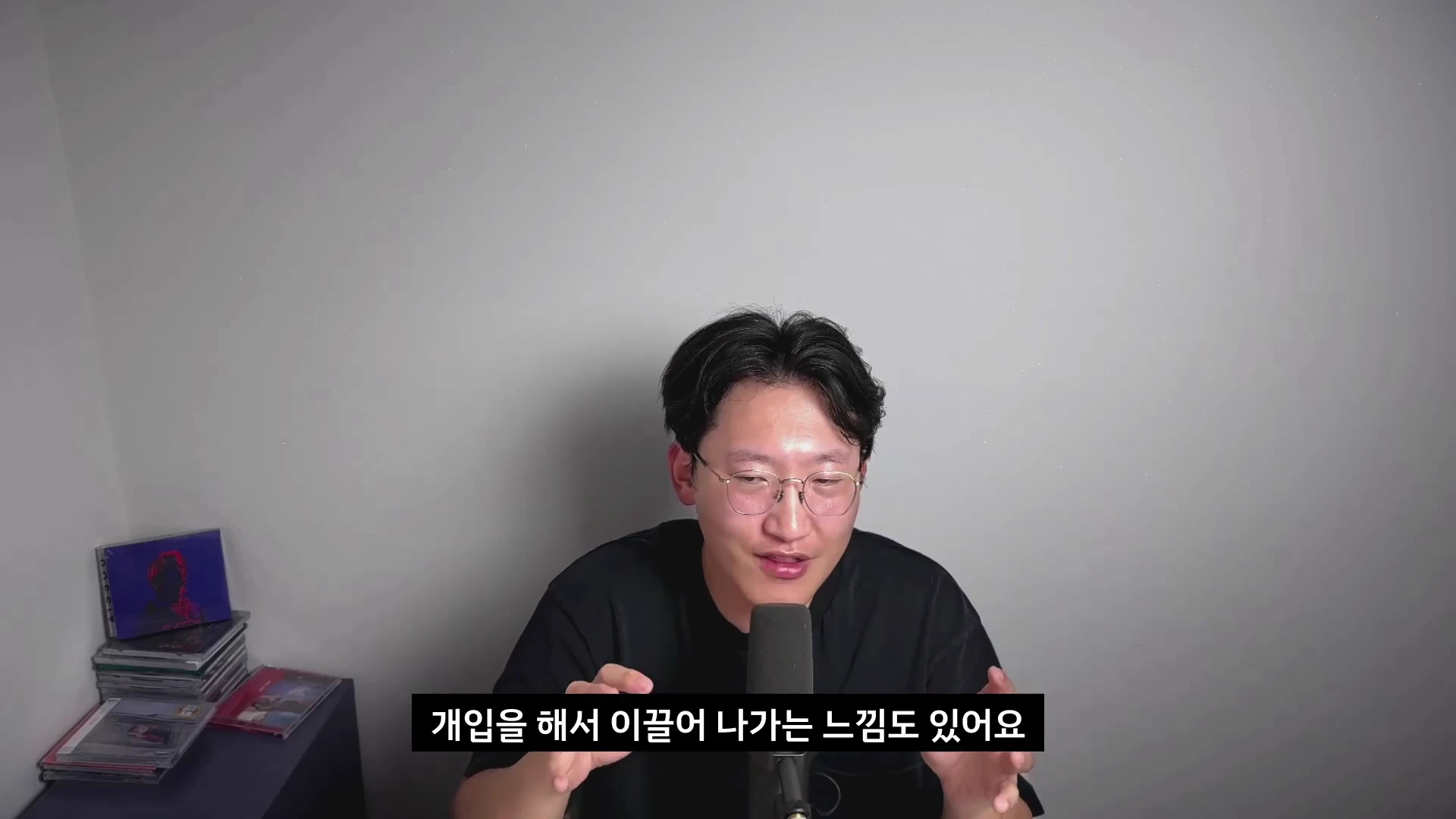 안경을 쓴 남성이 마이크 앞에서 손동작을 하며 대화를 나누고 있으며, 하단에는 검은색 자막 바가 삽입되어 있다.