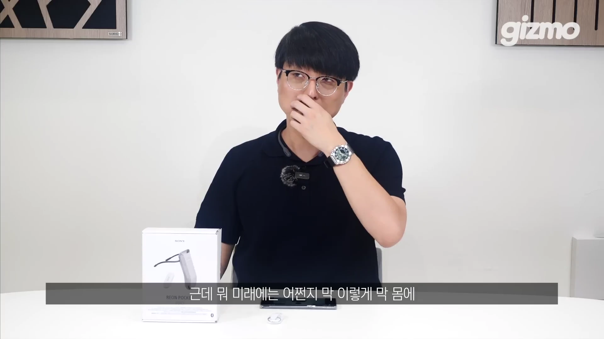 흰색 배경의 스튜디오에서 안경을 쓴 남성이 검은색 폴로셔츠를 입고 테이블 앞에 앉아 제품 패키지를 옆에 두고 생각에 잠긴 표정을 짓고 있다.