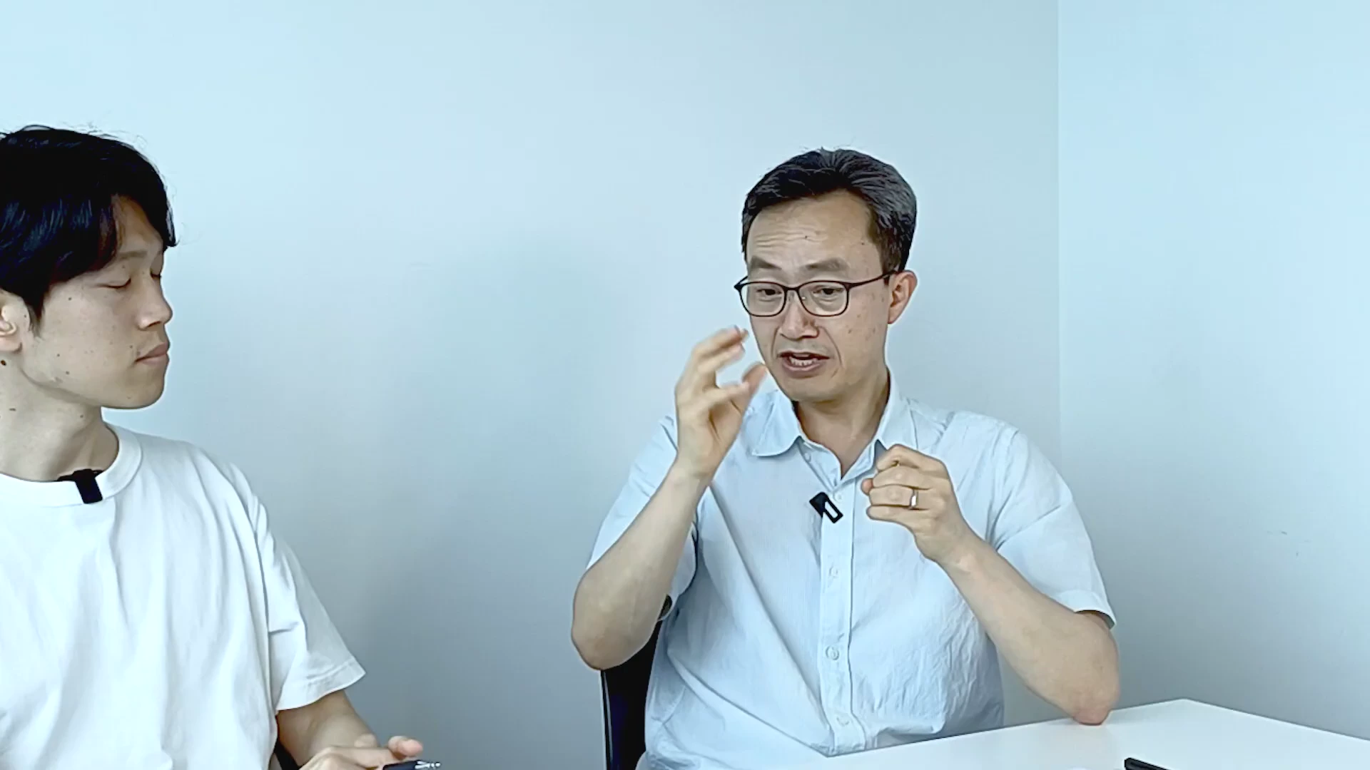 흰 배경의 스튜디오에서 흰 셔츠를 입은 안경 쓴 남성이 손짓하며 이야기하고 있고, 그 옆에 다른 남성이 앉아 경청하고 있는 모습이다.
