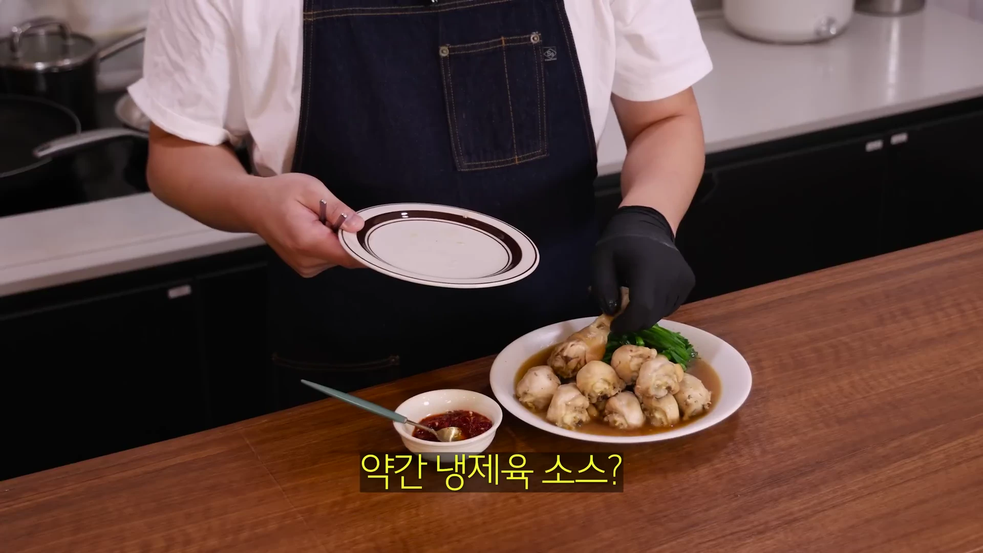 앞치마를 두르고 검은색 위생 장갑을 낀 사람이 완성된 닭다리 요리가 담긴 접시를 식탁 위에 놓고 있다.