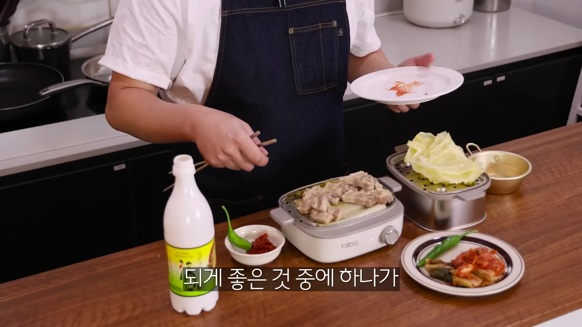 앞치마를 두른 요리사가 조리 기기에서 잘 익은 수육을 꺼내 접시에 담으려고 하며 식탁 위에는 막걸리와 김치가 차려져 있다.