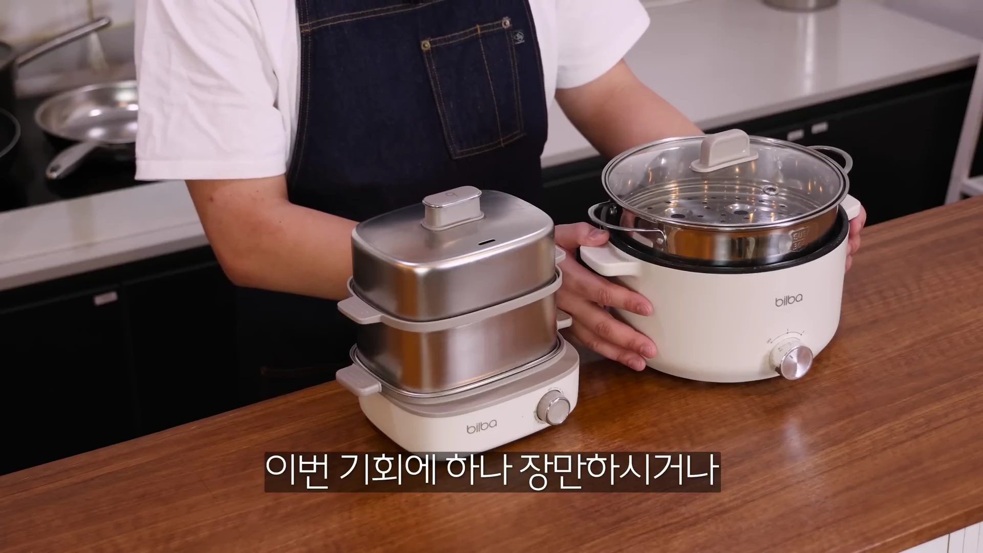 앞치마를 입은 사람이 주방 조리대 위에 놓인 두 개의 빌바 멀티쿠커를 양손으로 보여주고 있는 모습.
