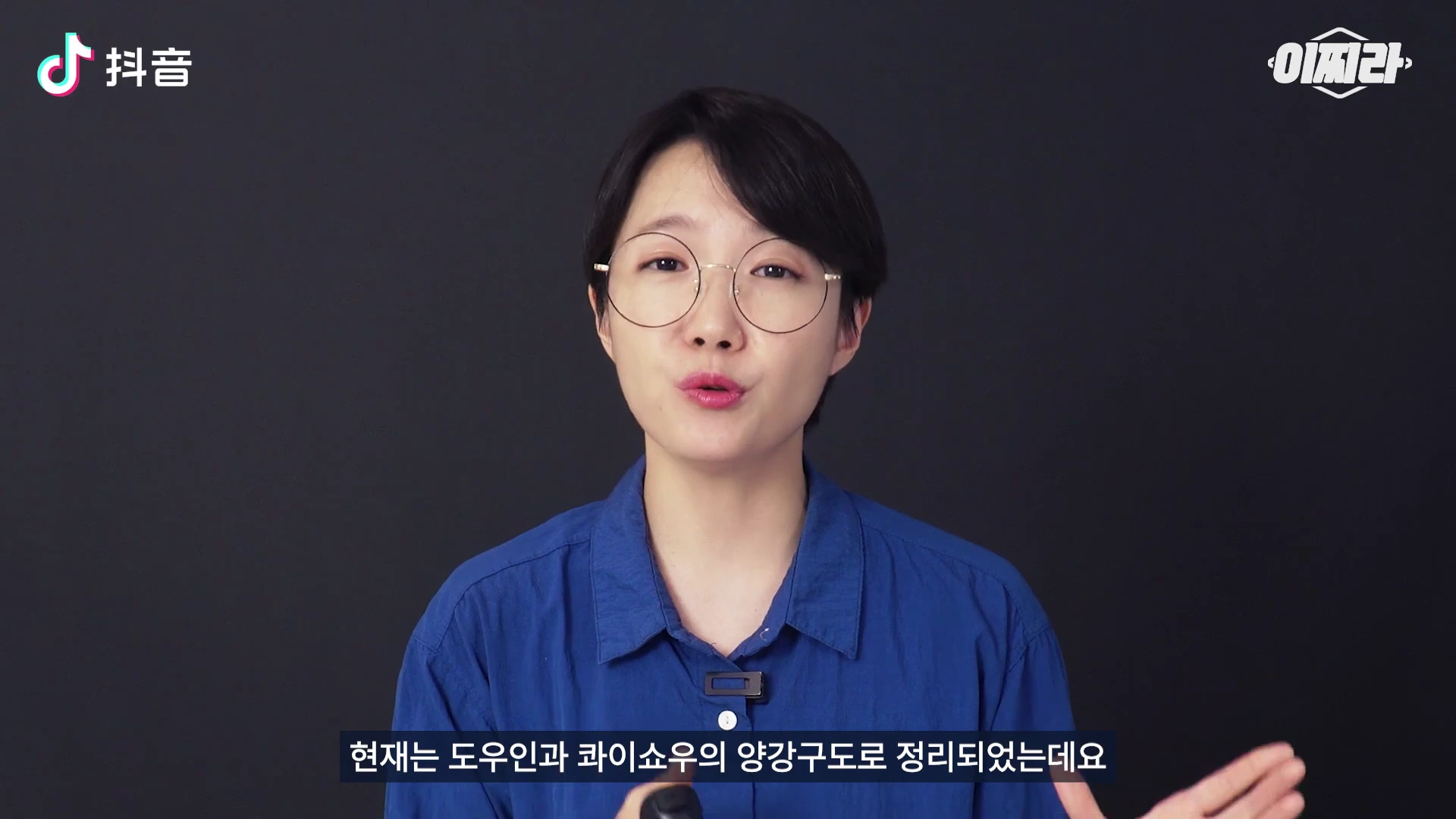 짧은 머리에 안경을 쓴 여성이 파란색 셔츠를 입고 검은색 배경 앞에서 무언가를 설명하고 있다. 화면 왼쪽 상단에는 중국어 텍스트 로고가 있고, 하단에는 한국어 자막이 표시되어 있다.