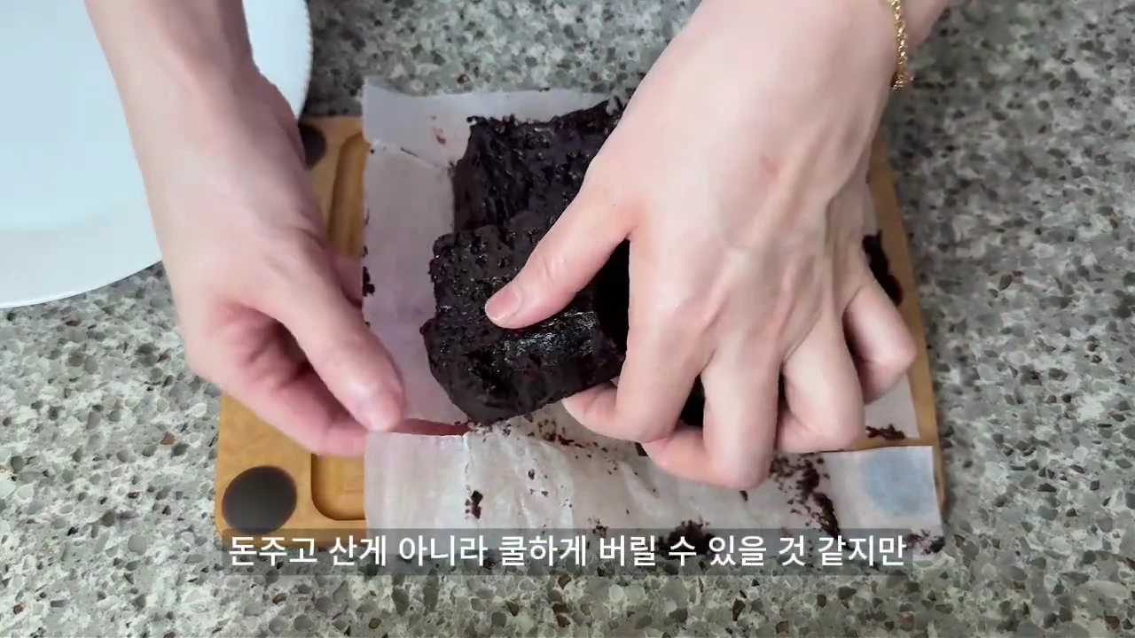 두 손으로 어두운색의 빵 조각을 종이 호일 위에서 조심스럽게 다루고 있는 모습과 하단에 한국어 자막이 적힌 영상 화면.