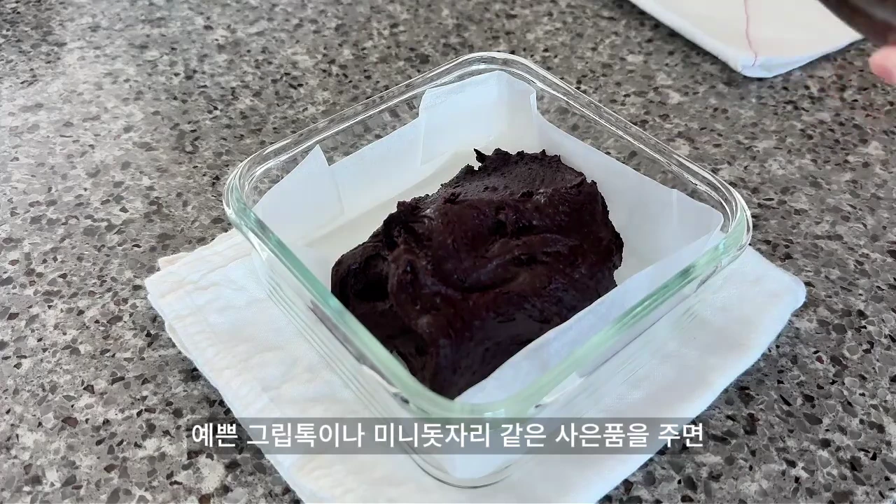 유리 밀폐 용기에 담긴 짙은 갈색의 반죽이 주방 대리석 조리대 위에 놓여 있으며, 화면 하단에는 한국어 자막이 표시되어 있습니다.