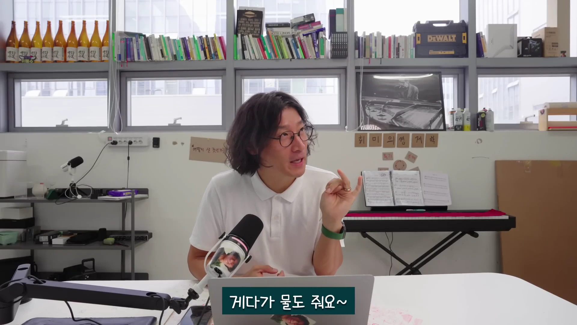 안경을 쓴 남성이 마이크 앞에서 손동작을 하며 이야기를 나누고 있는 모습이 담긴 영상 캡처 화면이며, 하단에는 '게다가 물도 줘요~'라는 한국어 자막이 적혀 있다.
