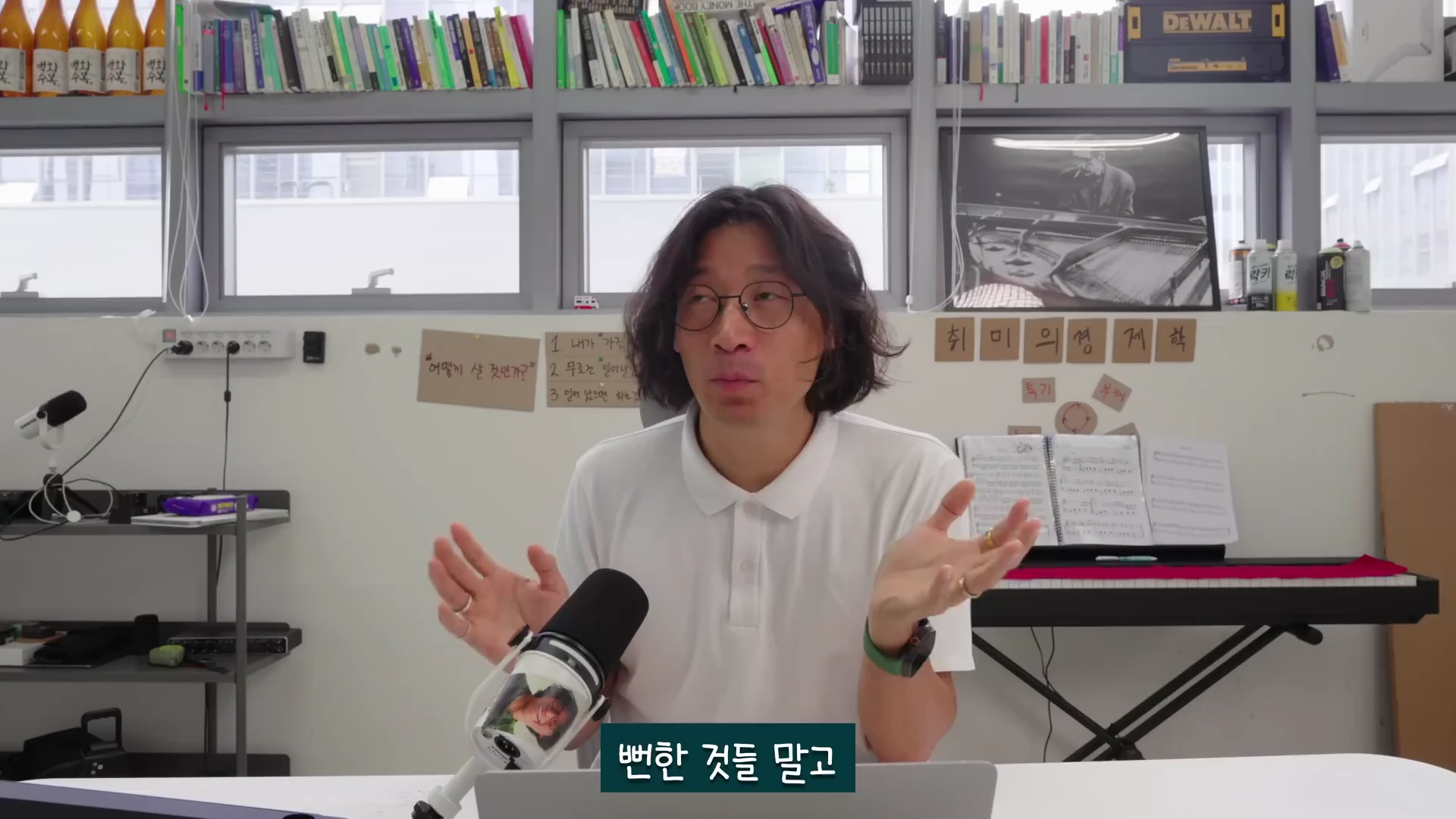 흰색 폴로셔츠를 입고 안경을 쓴 중단발 머리의 남성이 책상이 놓인 실내 공간에서 양손을 펼치며 이야기하는 영상 캡처 화면