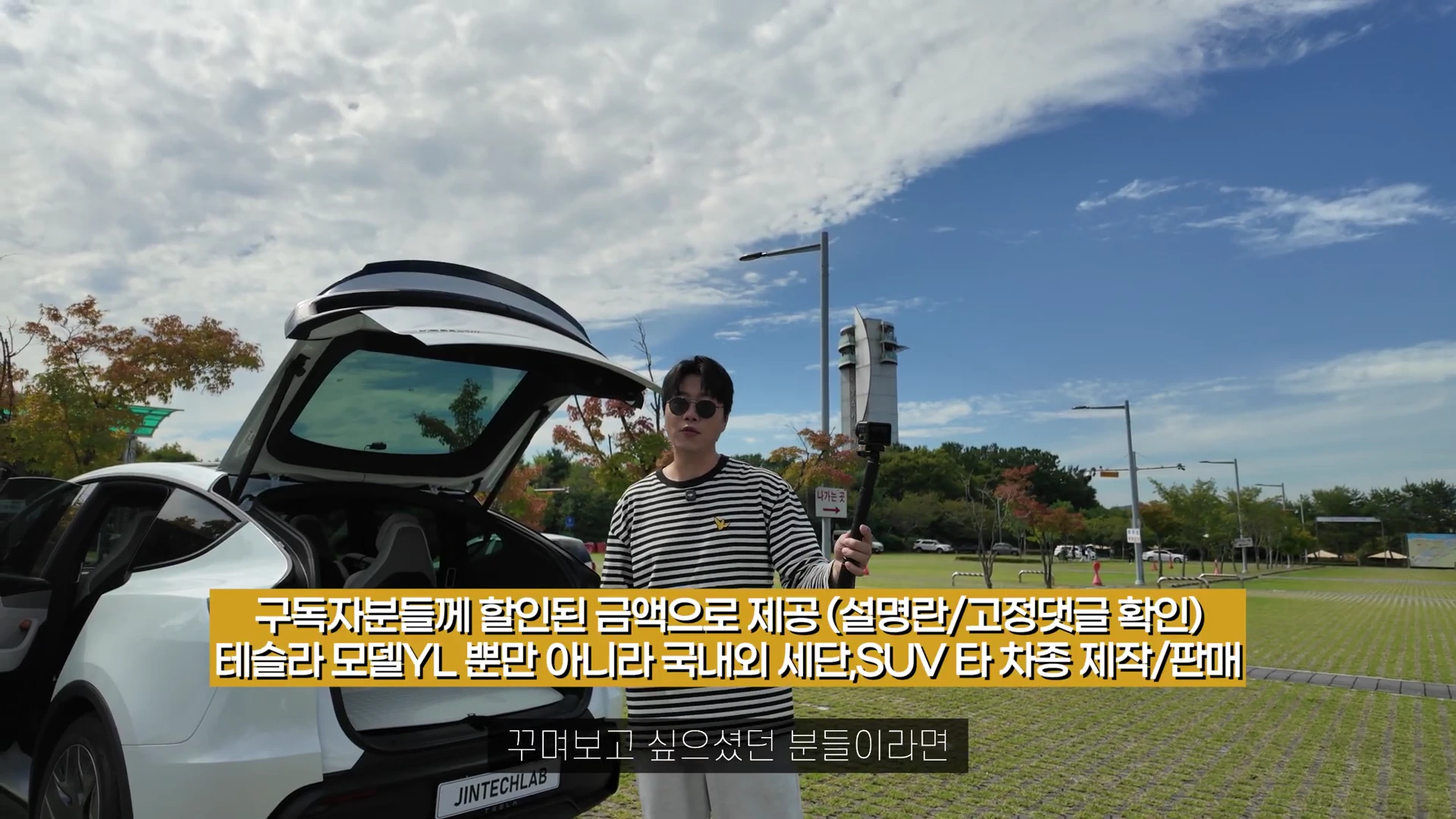 선글라스를 쓴 남성이 야외 주차장에서 트렁크가 열린 흰색 테슬라 차량 옆에 서서 카메라를 들고 서 있는 모습