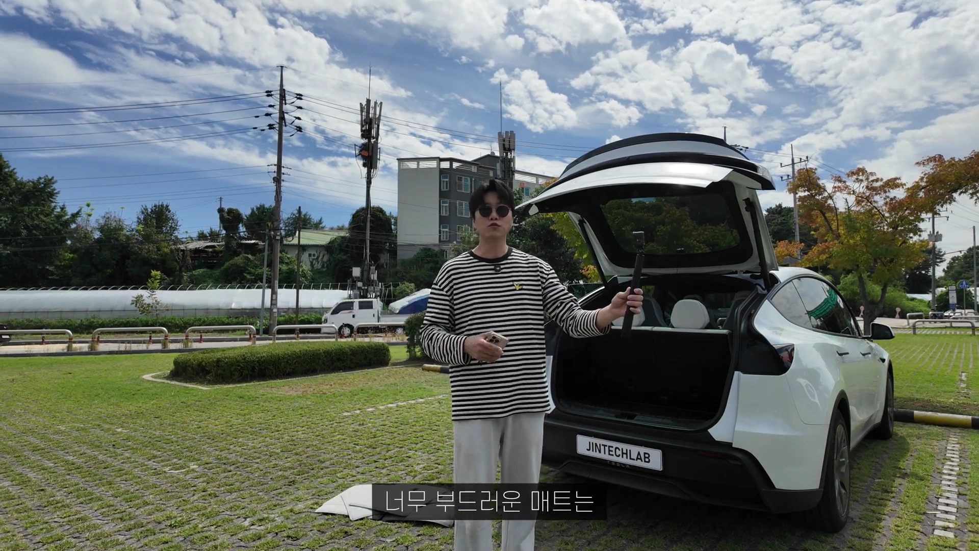 A man in a striped long-sleeved shirt and sunglasses stands outdoors in front of a white SUV with the trunk open, holding a device while gesturing during a video recording.