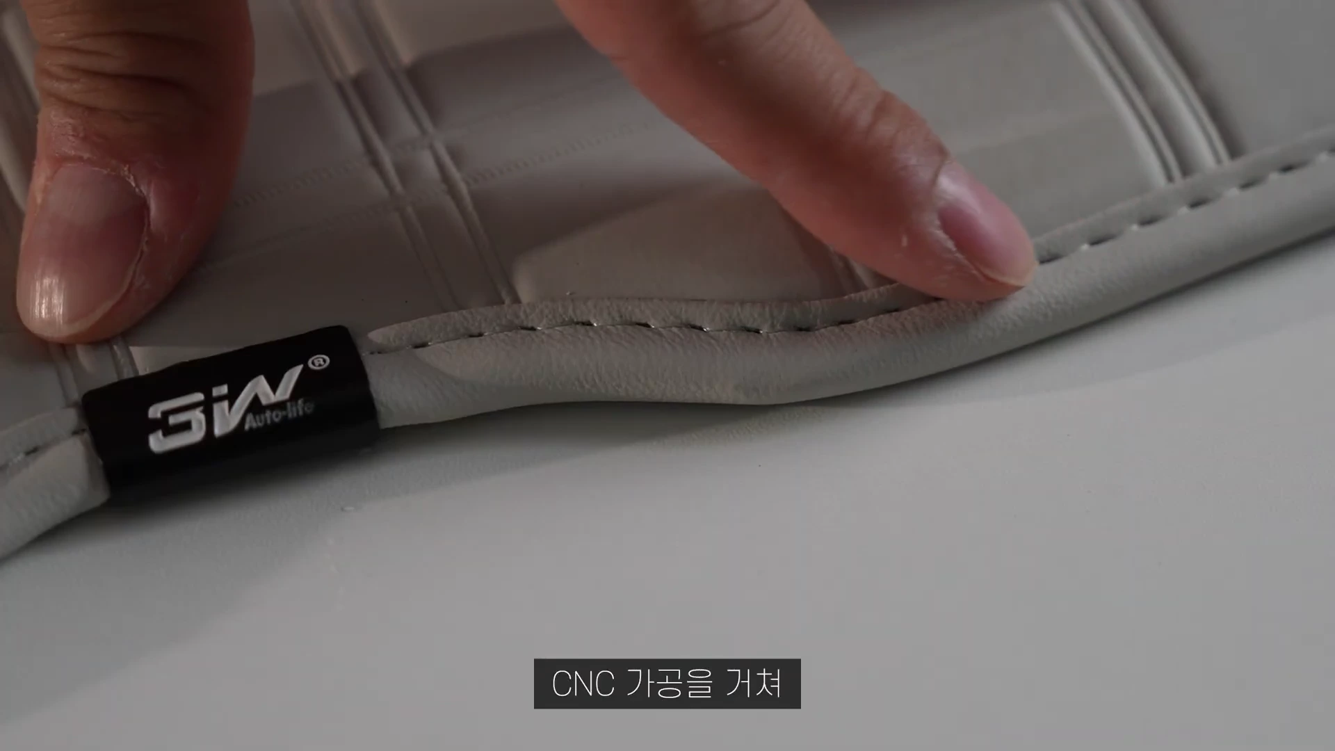 밝은 회색 가죽 매트의 테두리 부분을 손가락으로 가리키고 있는 모습으로 로고 태그와 촘촘한 박음질 처리가 보인다.