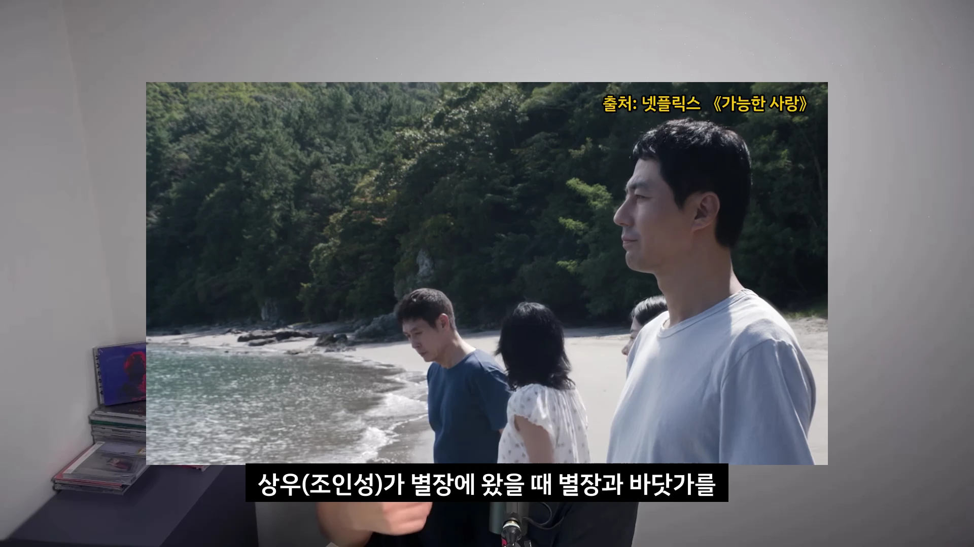 영화 속 장면으로 바닷가 모래사장에 서 있는 남녀 배우들의 모습이며 오른쪽에 서 있는 남성이 옆을 바라보고 있다.