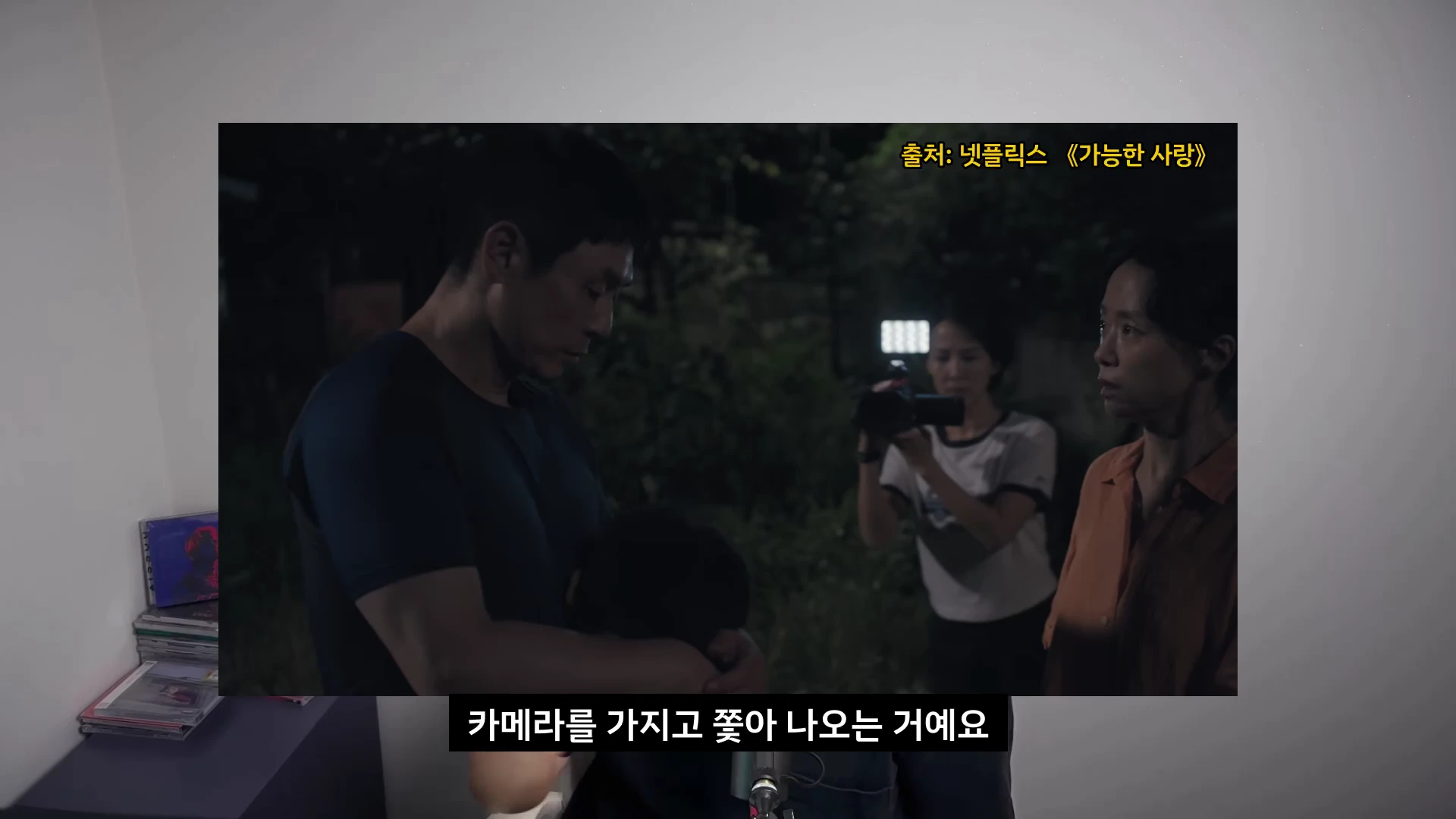 영화 속 어두운 밤, 한 여성이 카메라를 들고 다른 사람들을 촬영하는 장면이 담긴 스크린입니다.