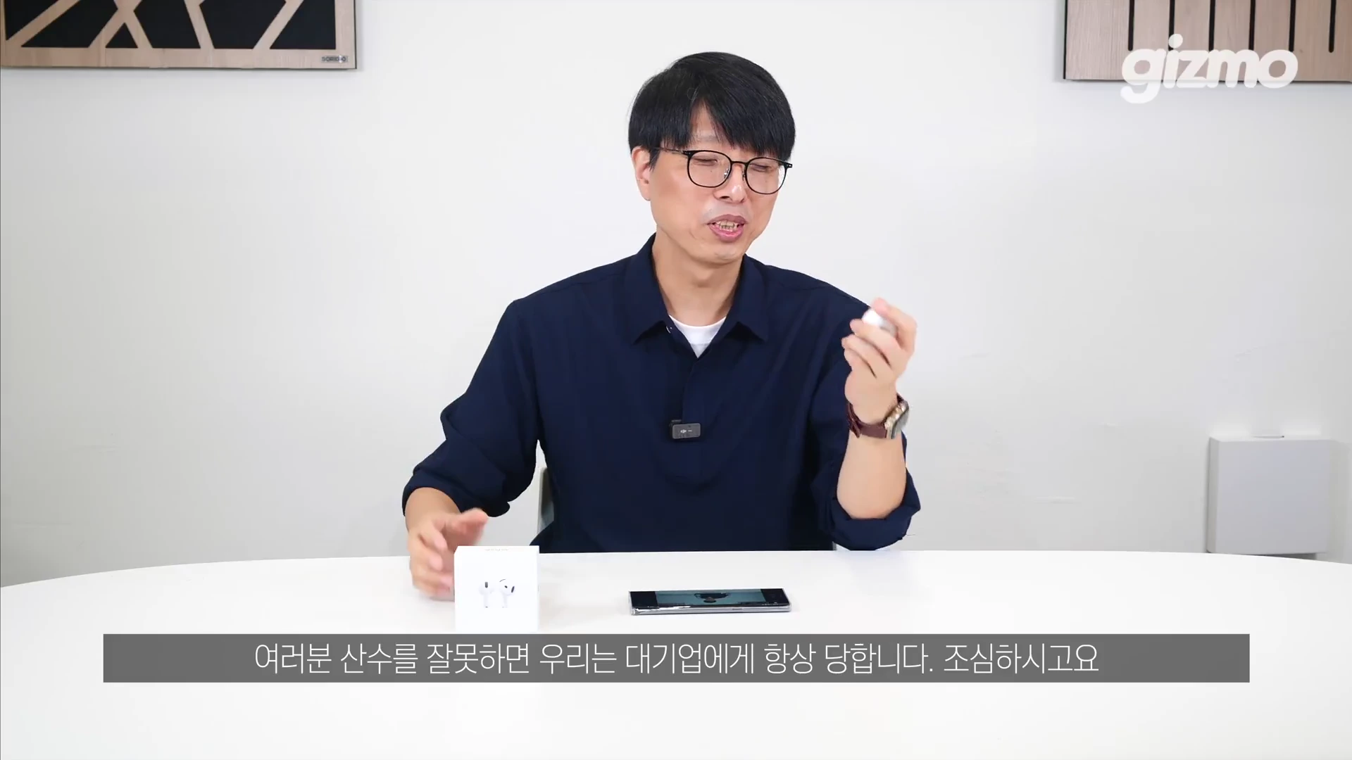 안경을 쓴 남성이 흰색 테이블에 앉아 손에 무선 이어폰을 들고 제품에 대해 설명하고 있는 스튜디오 인터뷰 영상의 한 장면입니다.
