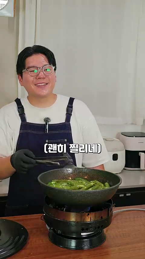 안경을 쓴 요리사가 앞치마를 두르고 핀셋으로 팬에 담긴 청양고추를 뒤집으며 밝게 웃고 있는 모습
