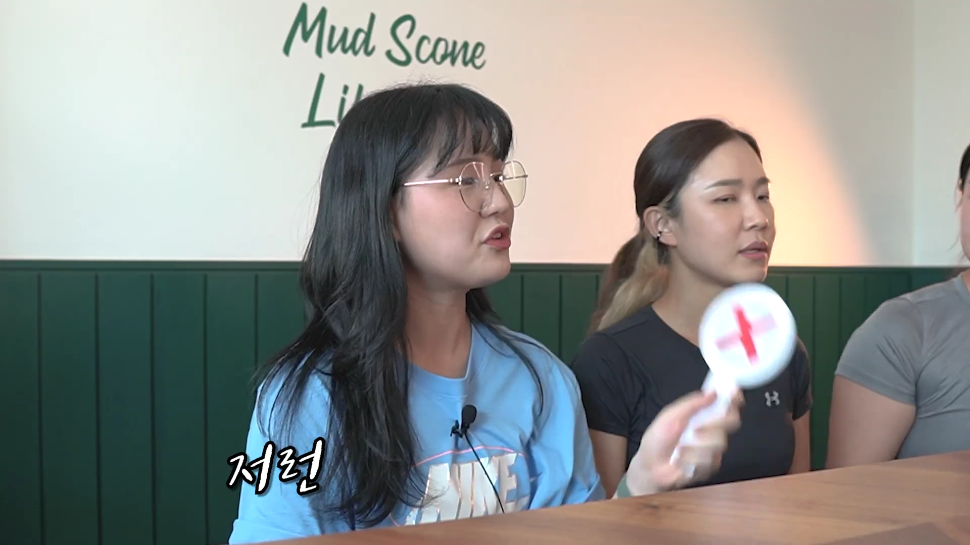 안경을 쓴 여성이 말하며 손에 빨간색 엑스(X) 표시가 그려진 팻말을 들고 있고, 옆에 다른 여성들이 앉아 있는 모습.