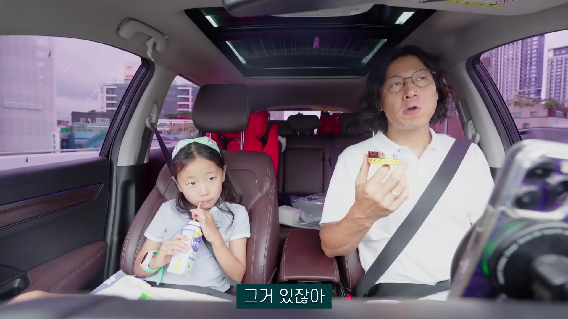 차 뒷좌석에 앉은 어린 딸과 운전석에 앉은 중년 남성이 서로 대화하고 있는 모습
