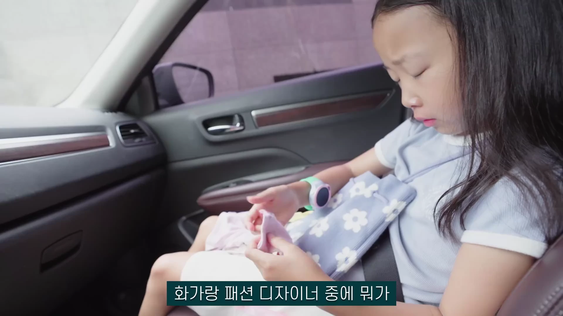 자동차 뒷좌석에 앉은 어린아이가 무릎 위에 놓인 작은 옷을 손으로 만지작거리며 아래를 내려다보고 있는 영상 캡처 화면.