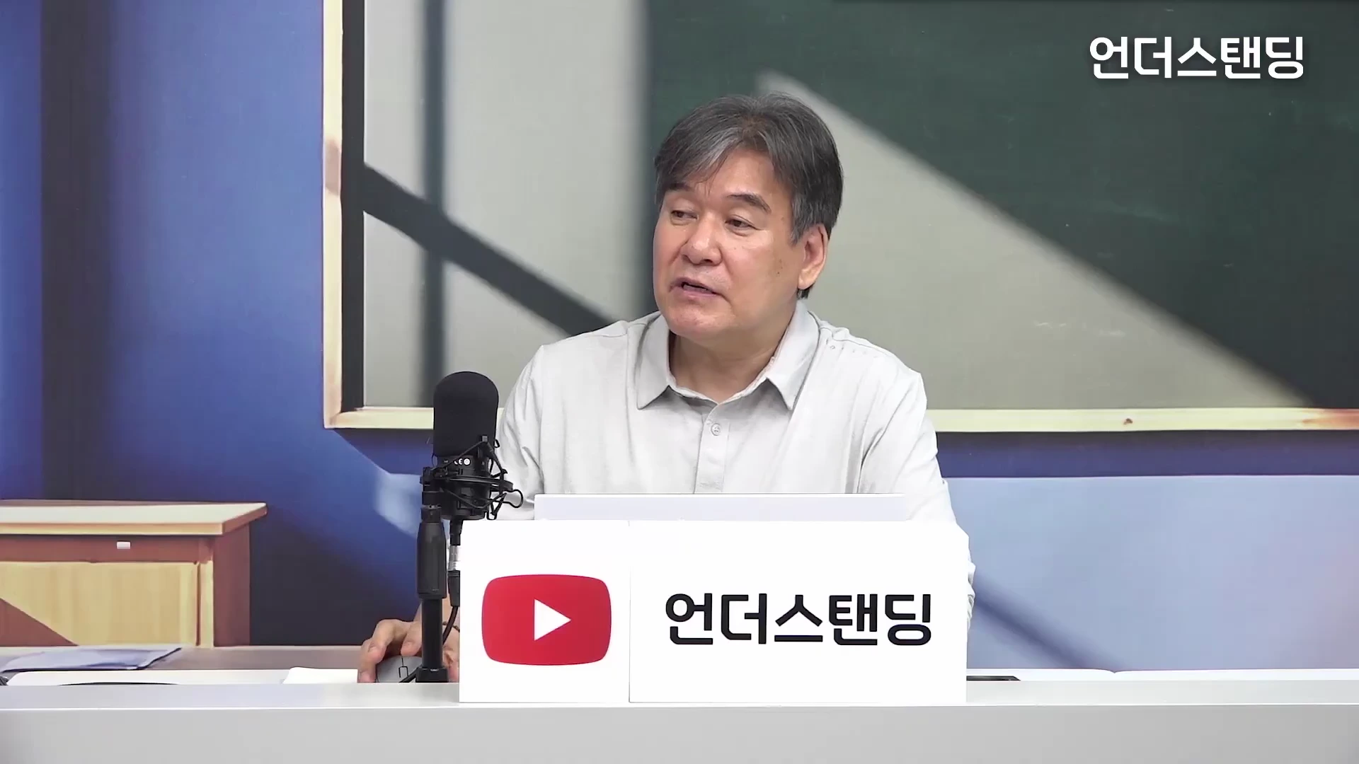스튜디오에 앉아 있는 중년 남성 전문가가 마이크 앞에서 이야기를 하고 있고, 앞에는 방송사 로고가 적힌 흰색 명판이 놓여 있습니다.