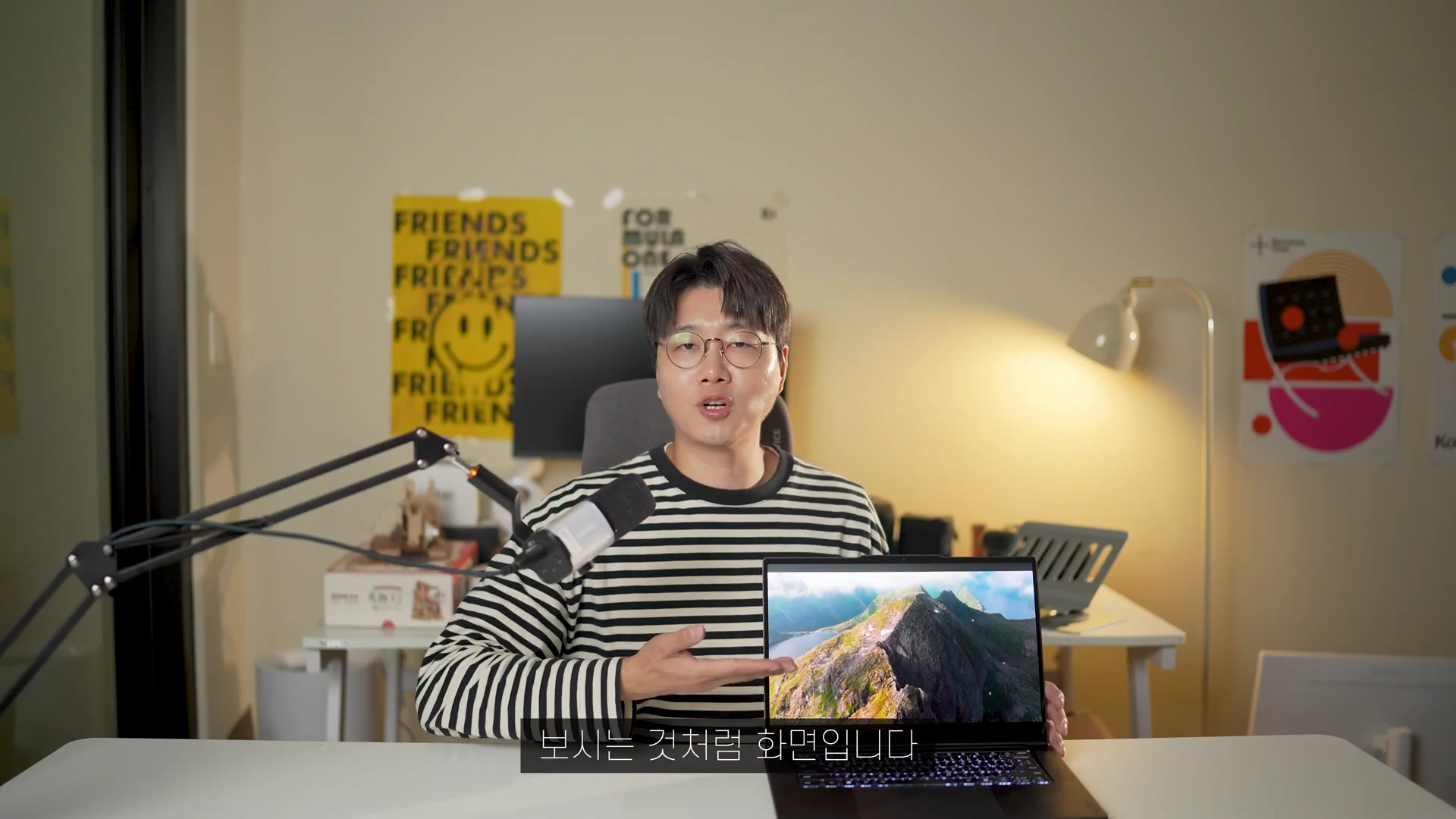 안경을 쓴 남성이 줄무늬 티셔츠를 입고 책상 앞에 앉아, 손으로 펼쳐진 노트북 화면을 가리키며 설명하고 있는 모습