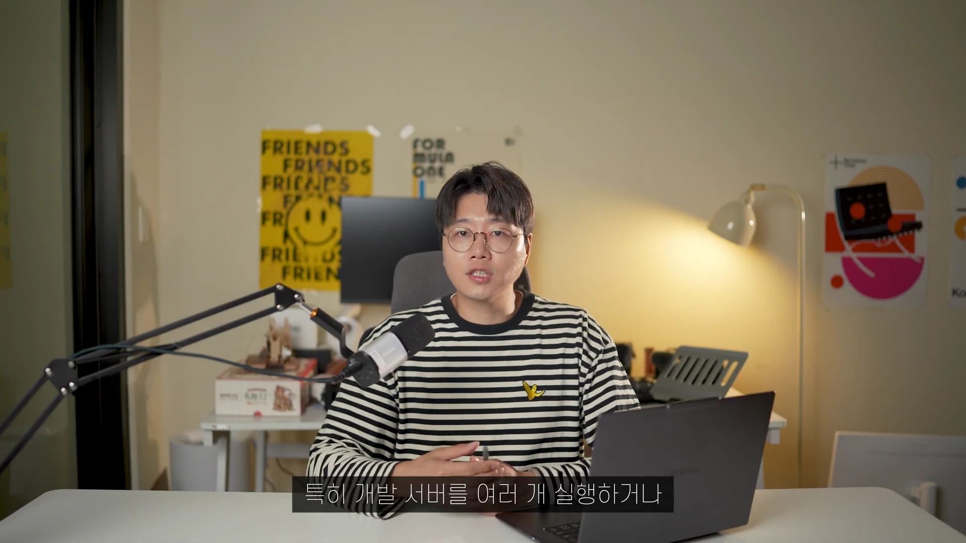 안경을 쓴 남성이 책상 앞에 앉아 마이크를 향해 이야기하고 있으며, 앞에는 노트북이 놓여 있고 뒤쪽 벽에는 노란색 포스터와 스탠드 조명이 보입니다.