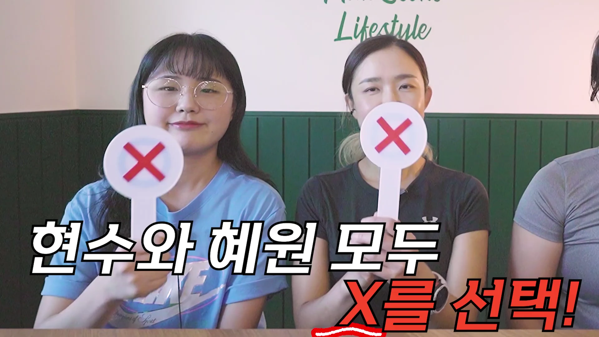 두 명의 여성이 카메라를 정면으로 바라보며 손에 빨간색 'X' 표시가 적힌 팻말을 들고 있는 모습.