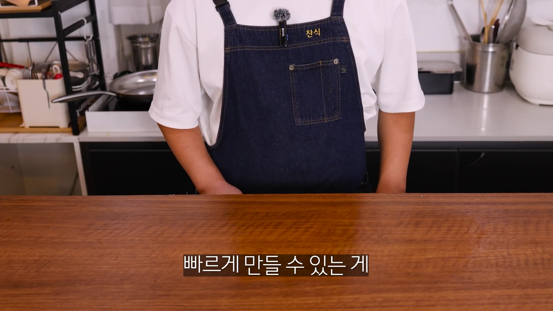 흰색 반소매 티셔츠 위에 남색 앞치마를 두른 사람이 부엌 조리대 앞에 서 있는 모습