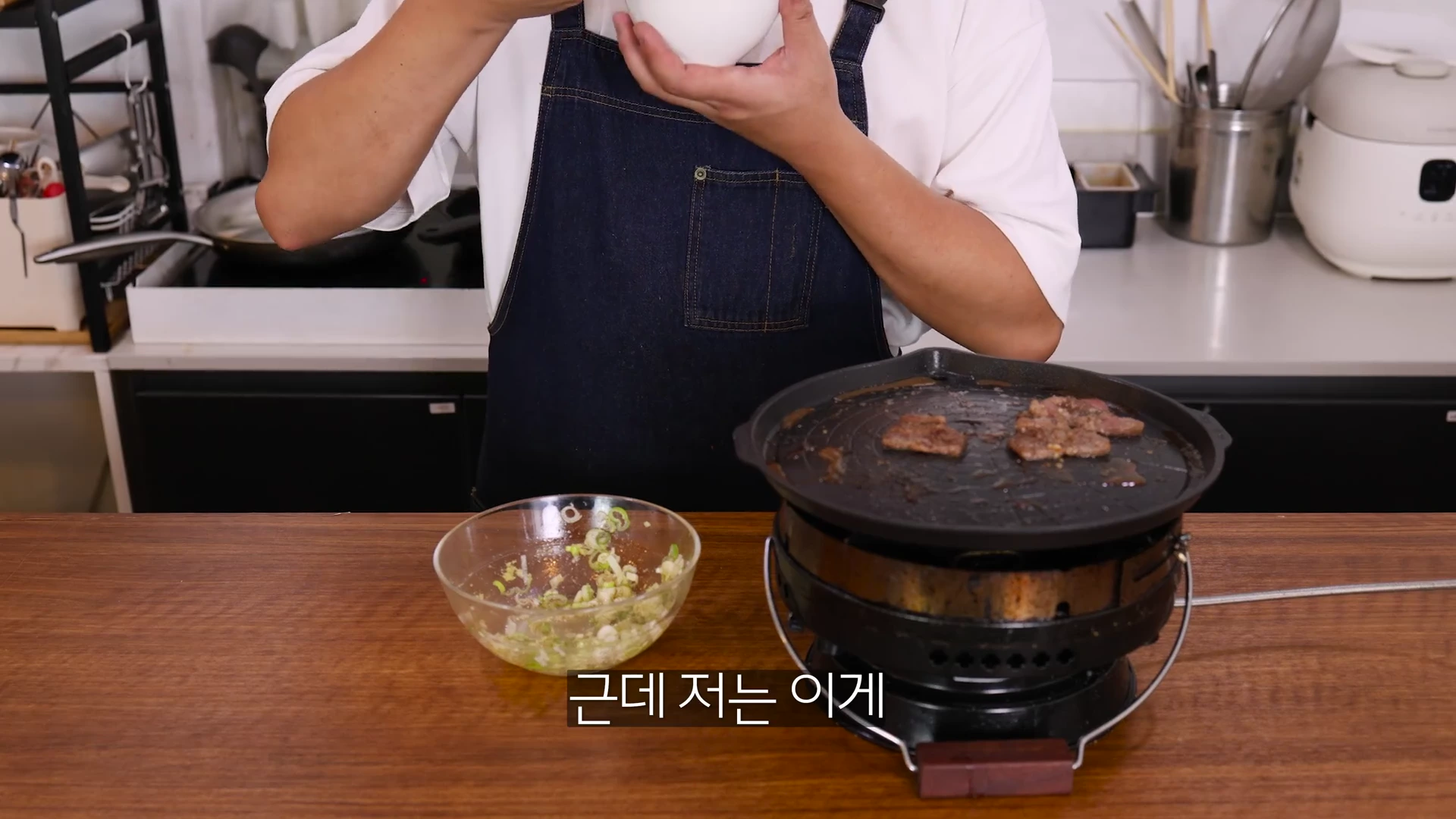 앞치마를 두른 요리사가 식탁 위에 놓인 화로와 파무침 그릇 앞에서 요리하고 있는 상반신 모습