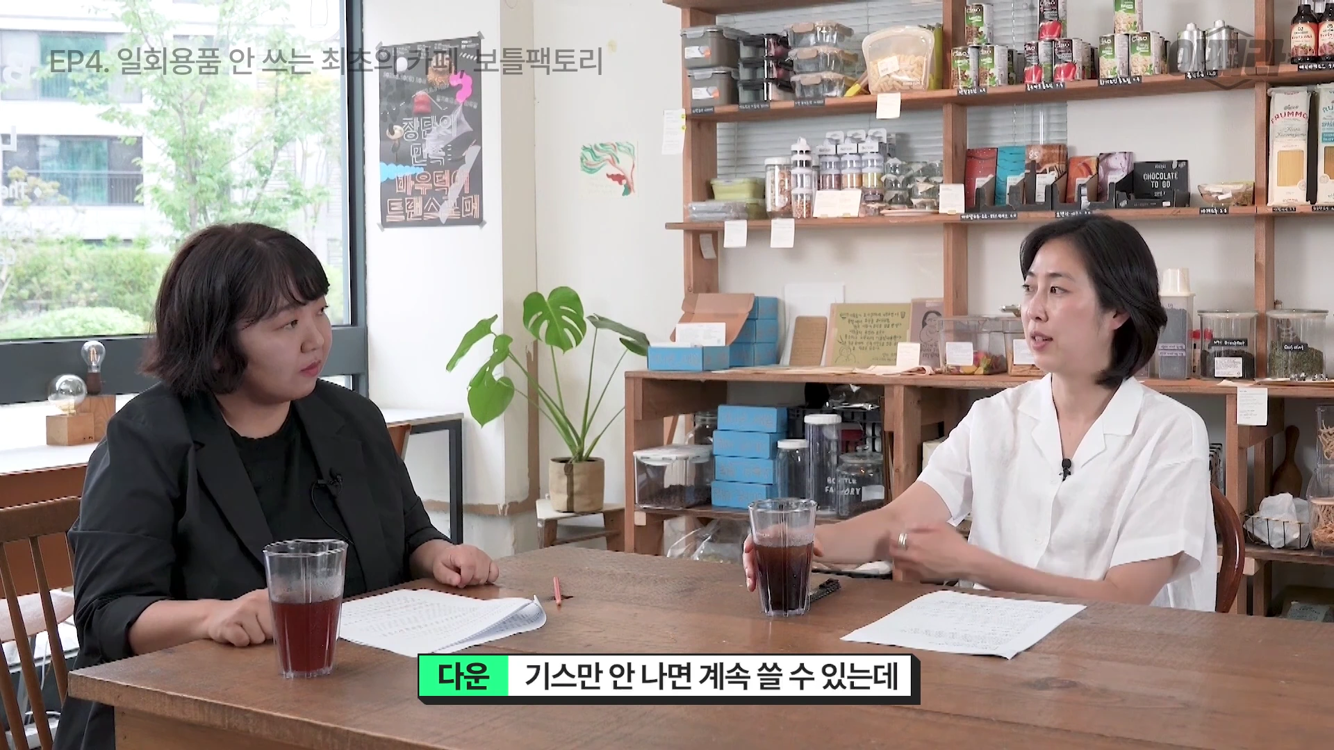 카페 테이블에 앉은 두 여성이 대화를 나누고 있으며, 앞에는 다회용 유리컵과 종이가 놓여 있고 뒤쪽에는 각종 식료품이 진열된 나무 선반이 보인다.