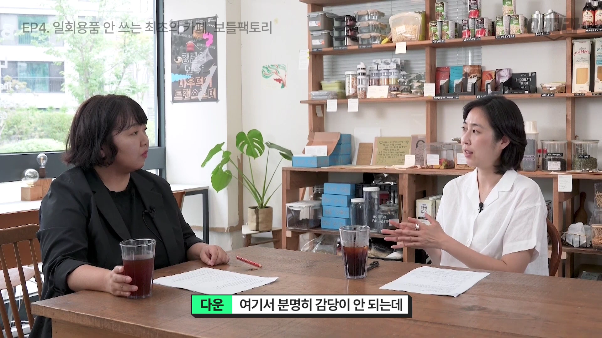 두 여성이 카페 내부 테이블에 앉아 서로 이야기를 나누고 있는 모습으로, 테이블 위에는 다회용 유리컵이 놓여 있고 배경에는 상품이 진열된 나무 선반이 보인다.