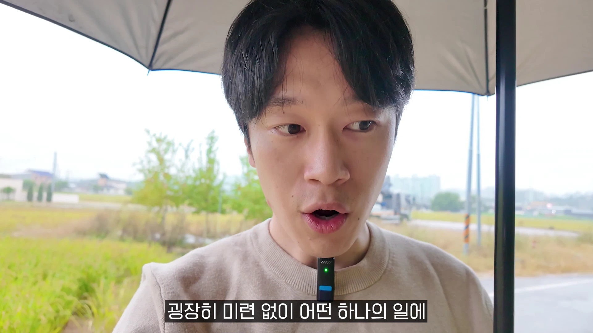 베이지색 니트를 입은 남성이 검은 우산을 쓰고 야외에서 이야기하고 있는 모습의 영상 캡처 화면이며, 화면 하단에는 자막이 적혀 있습니다.
