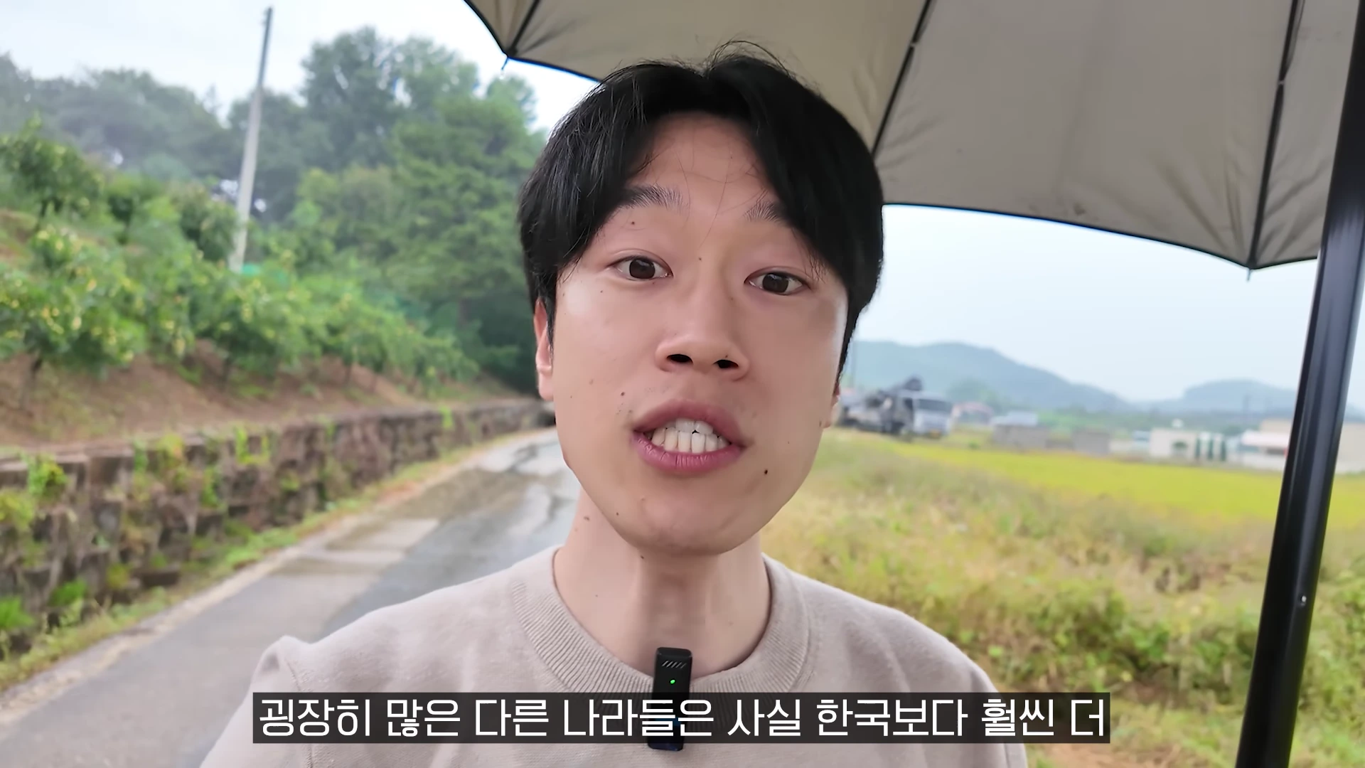 비 오는 야외에서 검은 우산을 쓰고 카메라를 보며 이야기하는 남성