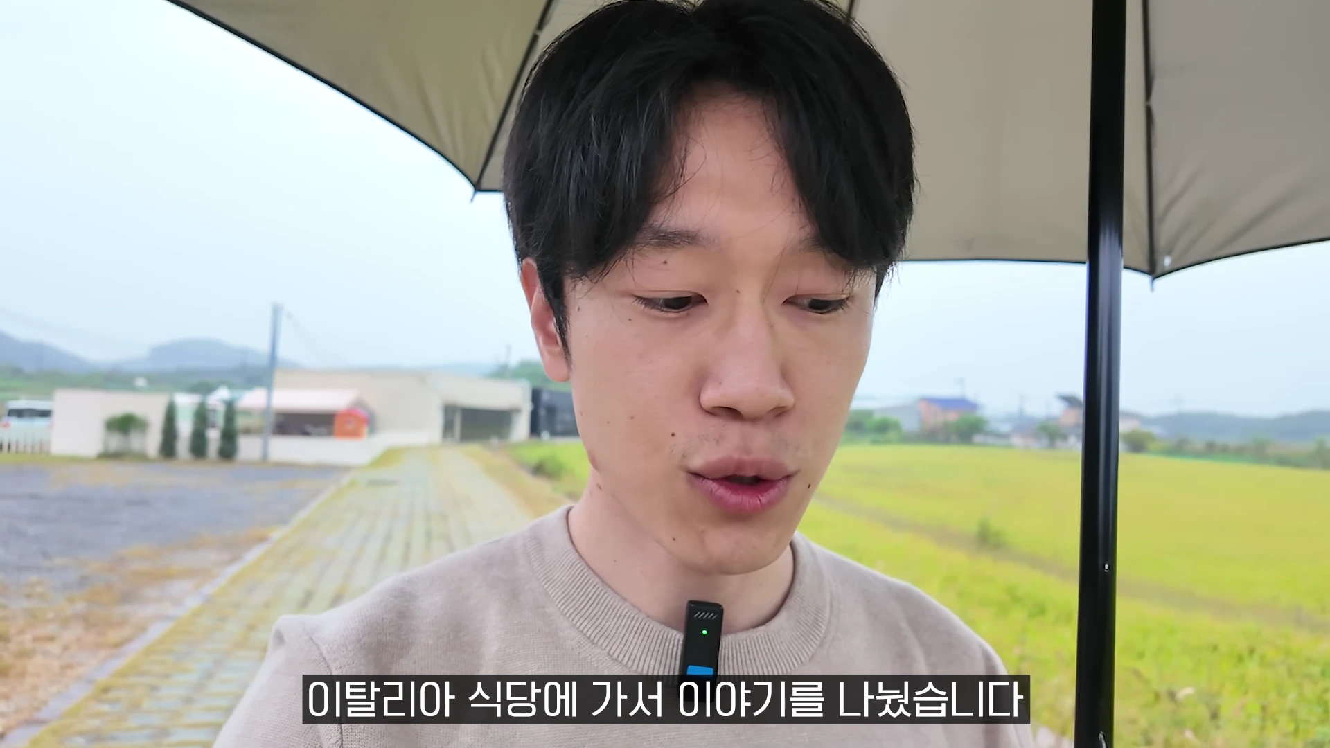 실외에서 검은색 우산을 쓰고 연한 베이지색 니트를 입은 남성이 정면을 바라보며 이야기를 하고 있는 모습입니다.