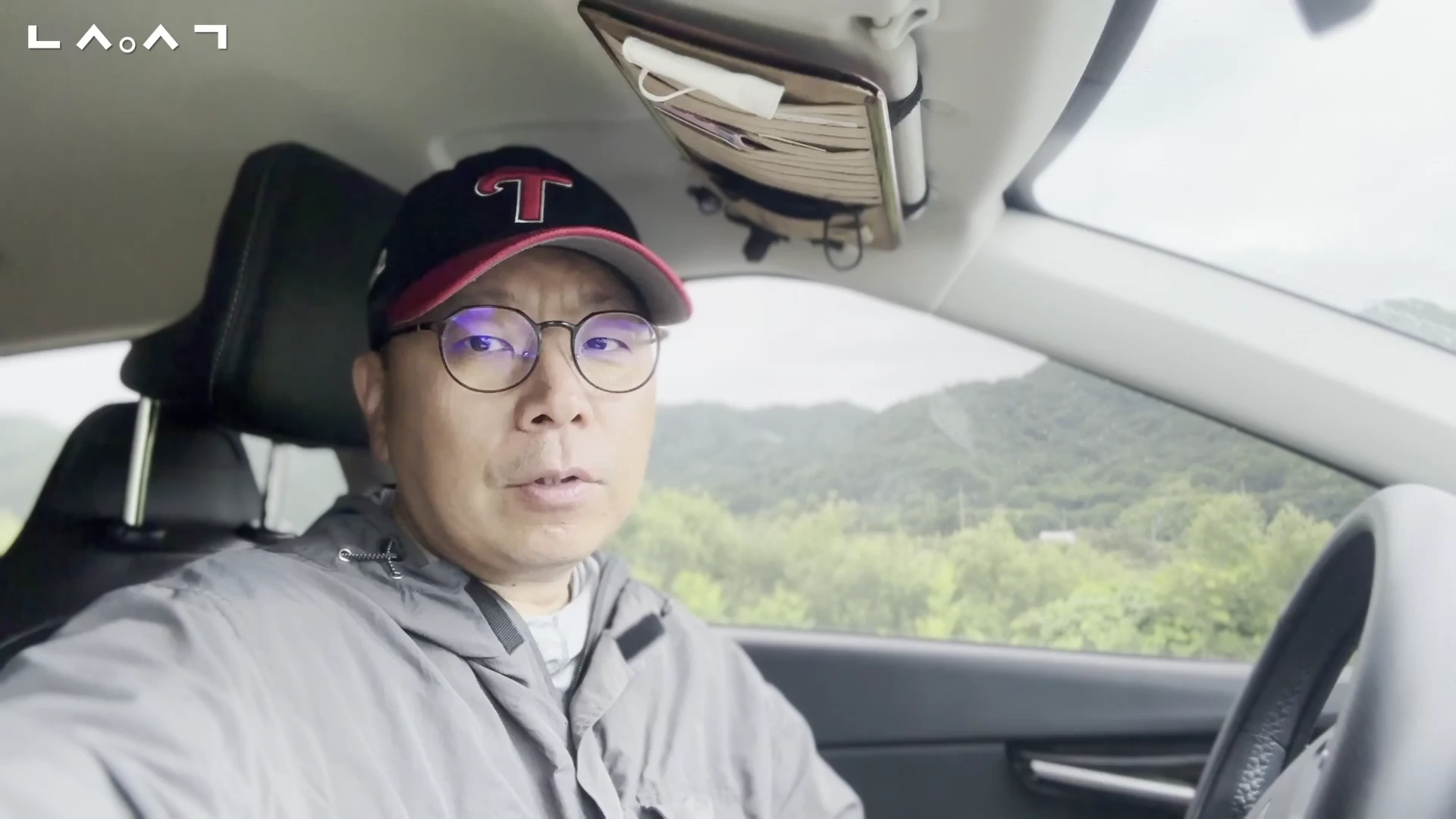 자동차 운전석에 앉아 카메라를 응시하며 이야기하는 중년 남성의 모습