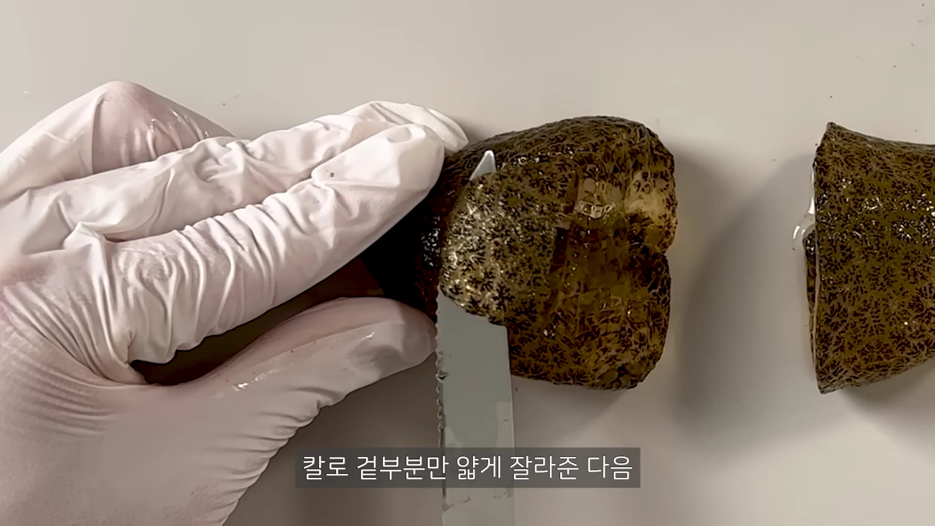 흰색 장갑을 낀 손이 작은 톱날 칼을 사용하여 갈색의 젤리 같은 덩어리를 얇게 잘라내는 모습이 담긴 근접 사진입니다.