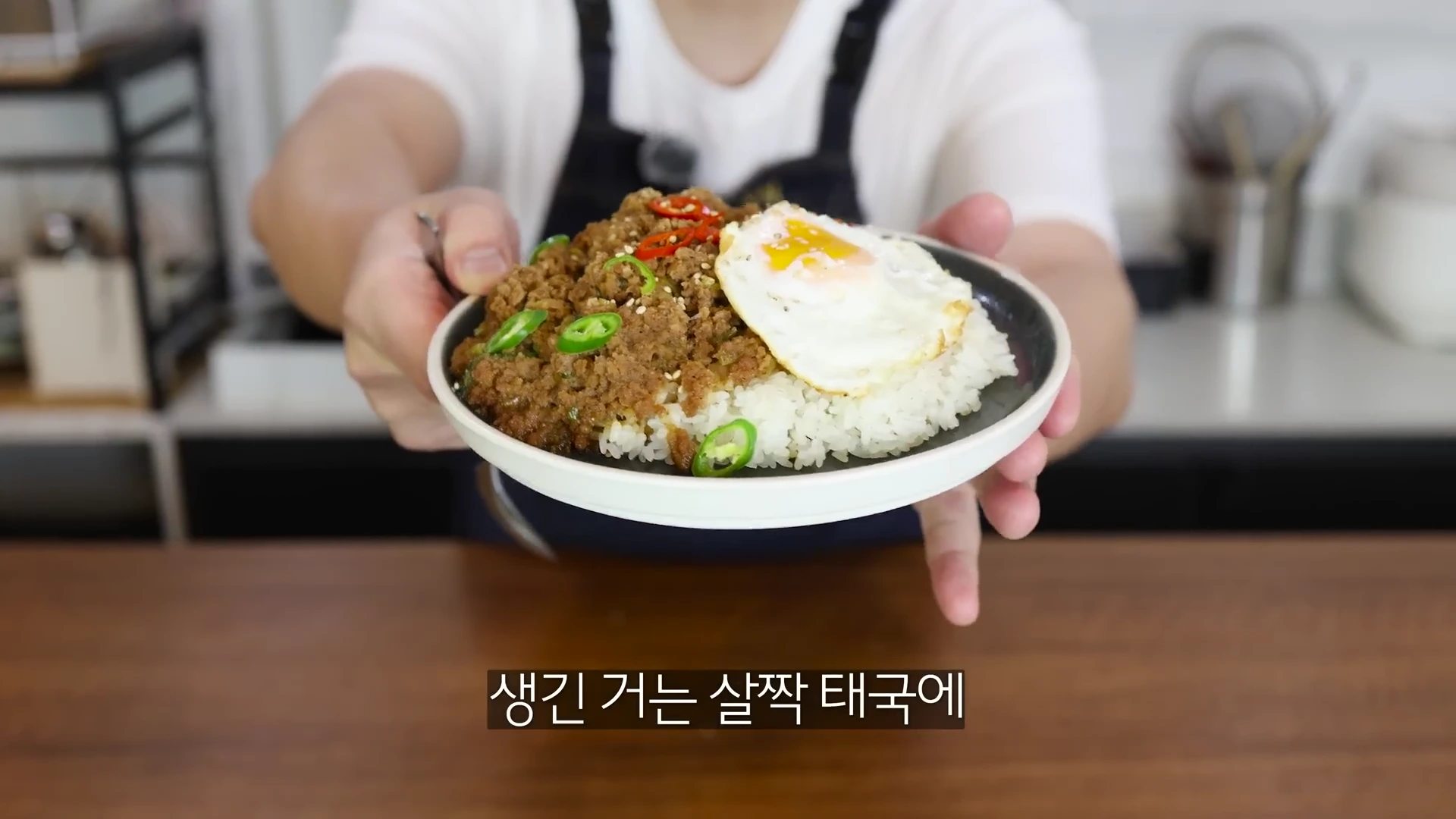 흰 티셔츠에 앞치마를 입은 사람이 다진 불고기, 계란 프라이, 고추가 올라간 밥 한 접시를 양손으로 들어 카메라 쪽으로 내밀고 있는 모습.