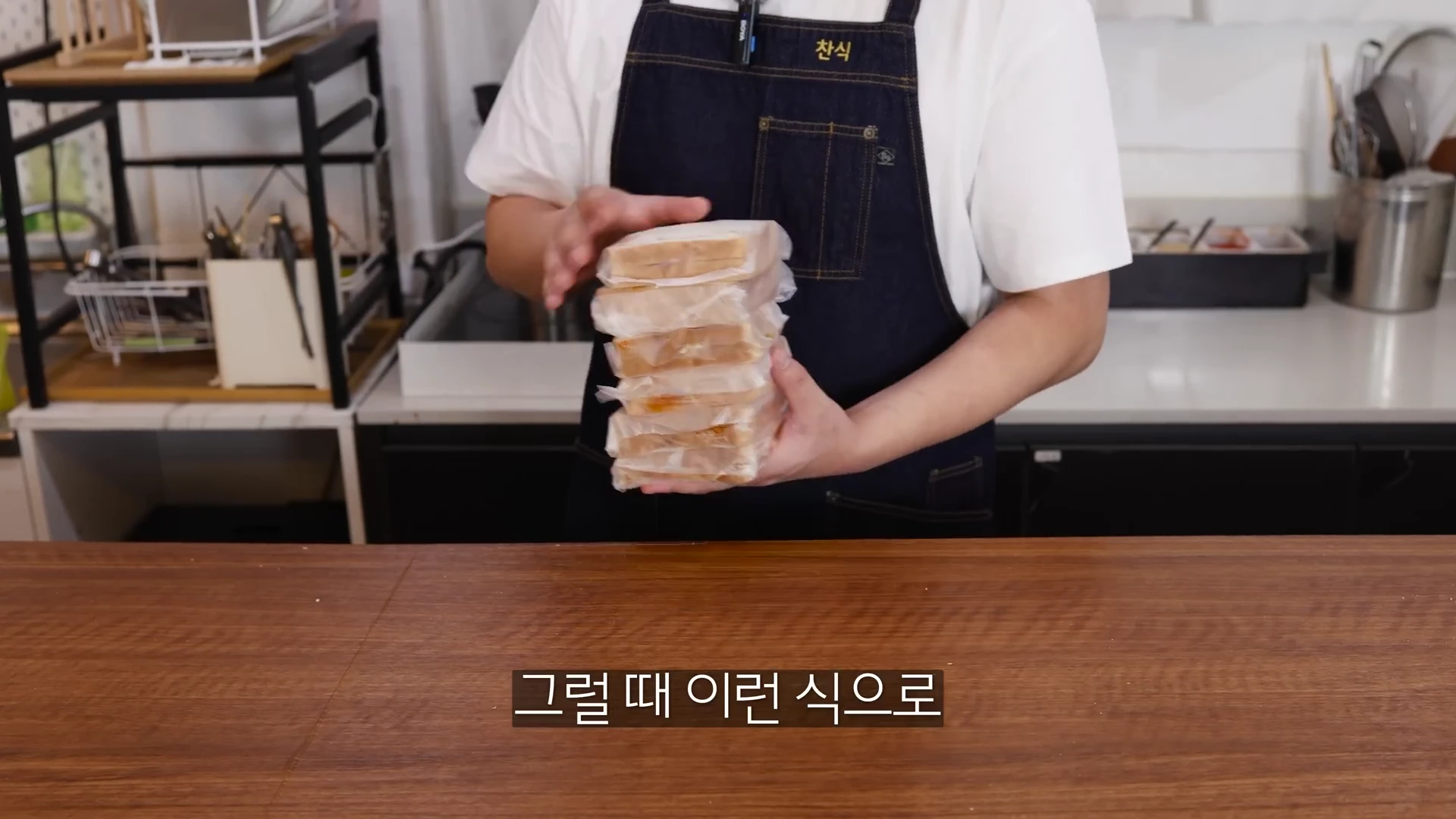 앞치마를 입은 사람이 비닐로 개별 포장된 식빵 샌드위치 여러 개를 쌓아 들고 있는 모습