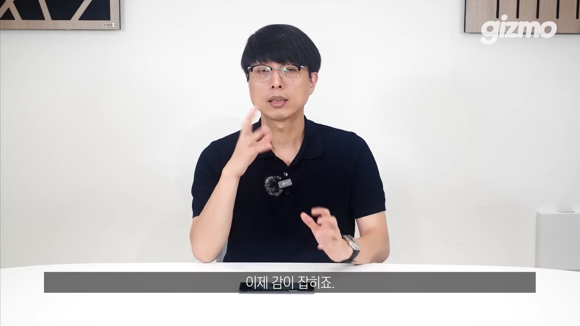 흰색 배경의 스튜디오에서 안경을 쓴 남성이 손짓을 하며 무언가를 설명하고 있는 모습