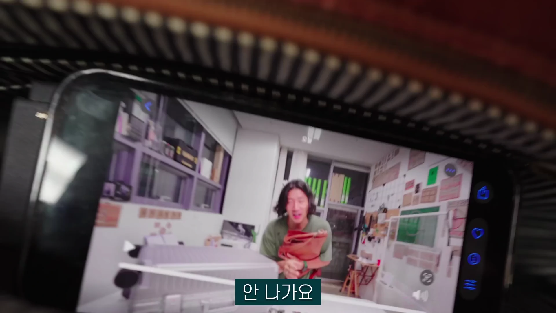실내에서 가방을 들고 서 있는 긴 머리의 남성이 스마트폰 화면에 재생되고 있으며, 화면 하단에는 '안 나가요'라는 자막이 표시되어 있다.