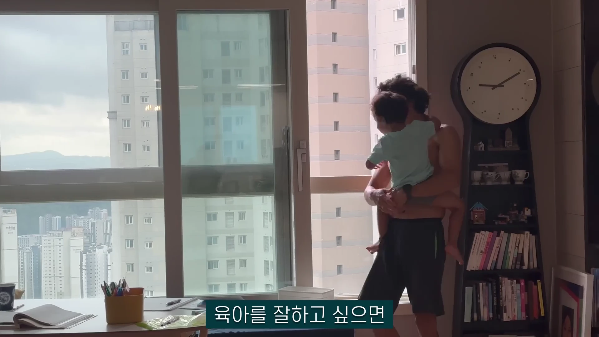 실내에서 한 남성이 아이를 안고 창밖을 바라보고 있으며, 옆에는 책장과 시계가 놓여 있고 화면 하단에는 '육아를 잘하고 싶으면'이라는 자막이 떠 있습니다.