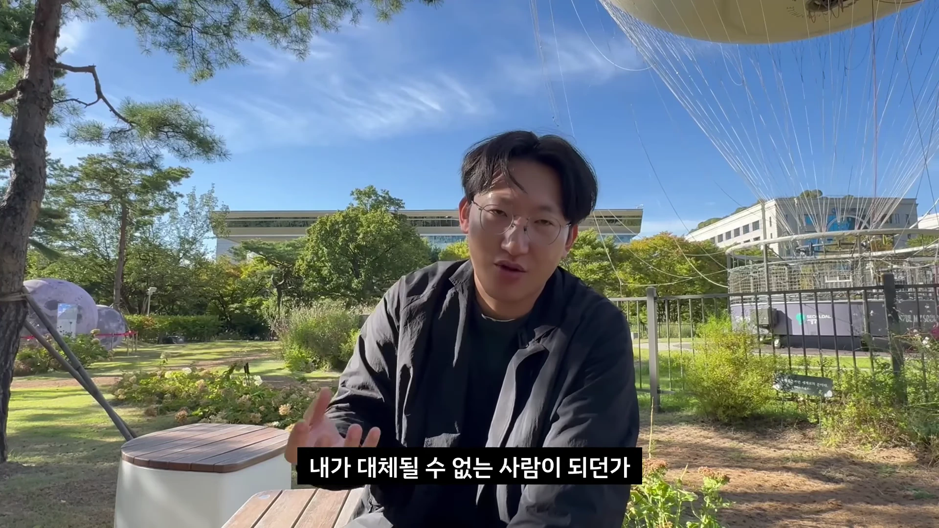 안경을 쓴 남성이 야외 공원에서 대화하며 손짓을 하고 있는 모습의 영상 화면입니다.