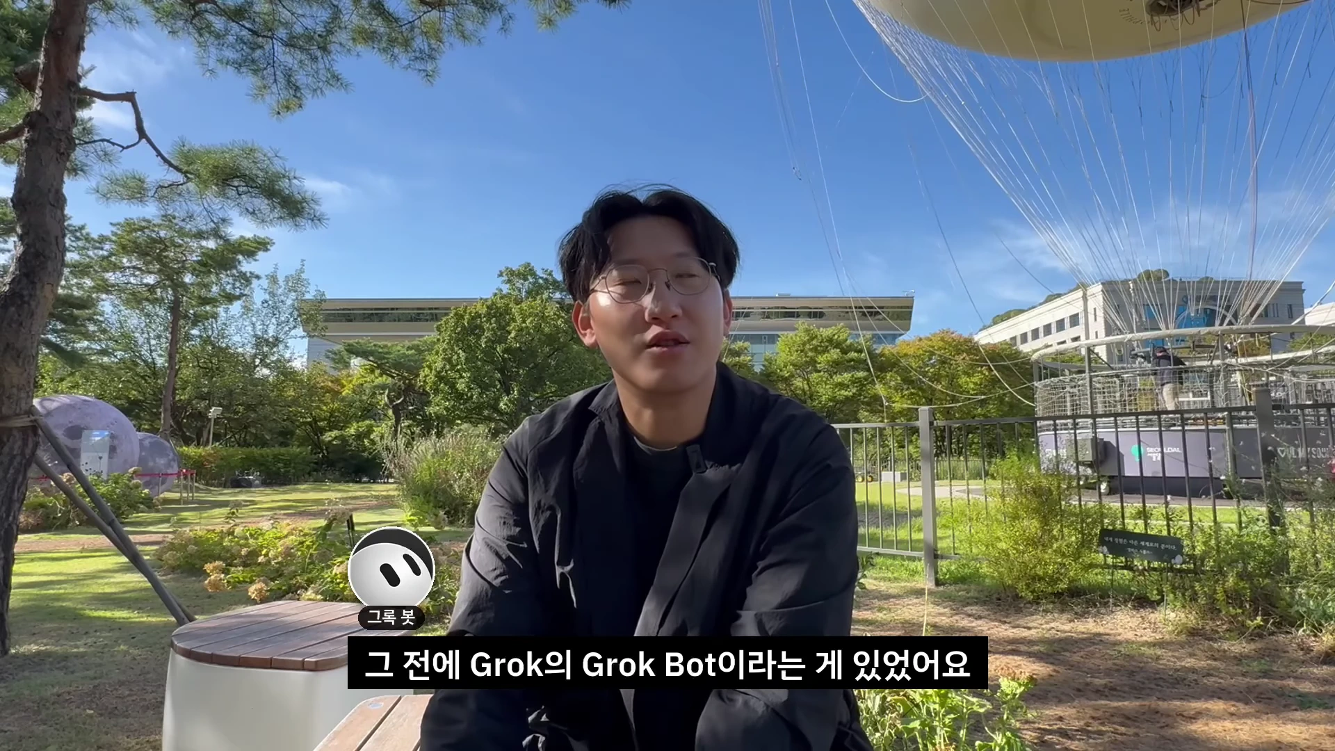 공원 벤치에 앉아 이야기하는 안경 쓴 남성, 화면 하단에는 자막과 그록 봇 아이콘이 표시된 영상 프레임