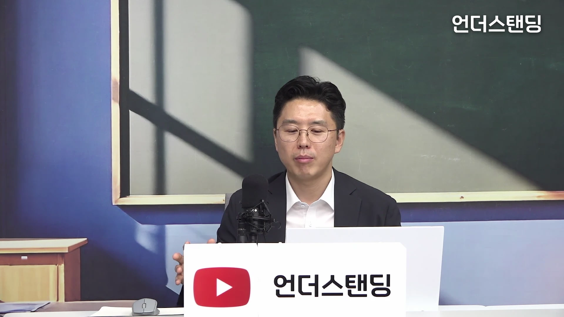 안경을 쓴 남성이 마이크 앞에 앉아 인터뷰를 진행하고 있으며 배경에는 칠판이 있는 스튜디오 공간이 보인다.