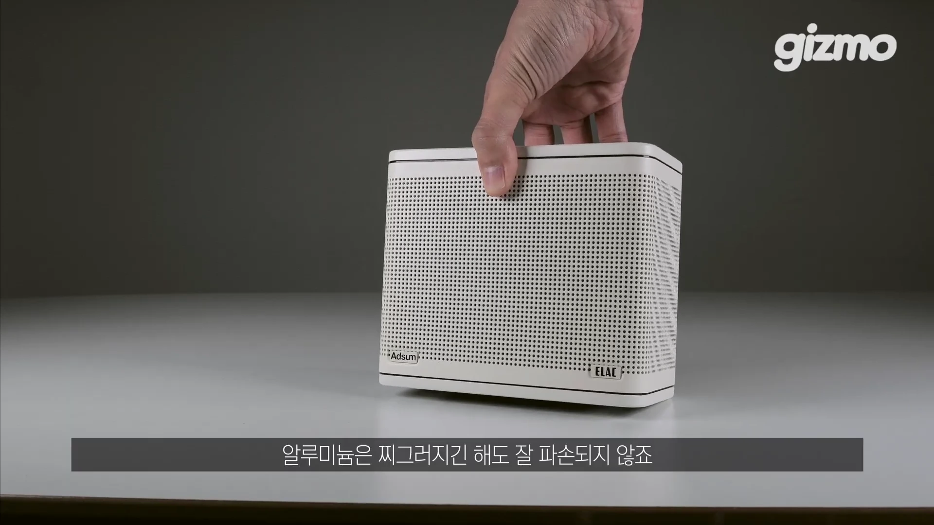 손으로 크림색 박스형 스피커를 들고 있는 모습. 전면에는 작은 도트 패턴 그릴과 왼쪽 하단에 애드섬 로고, 오른쪽 하단에 엘락 로고가 위치함.
