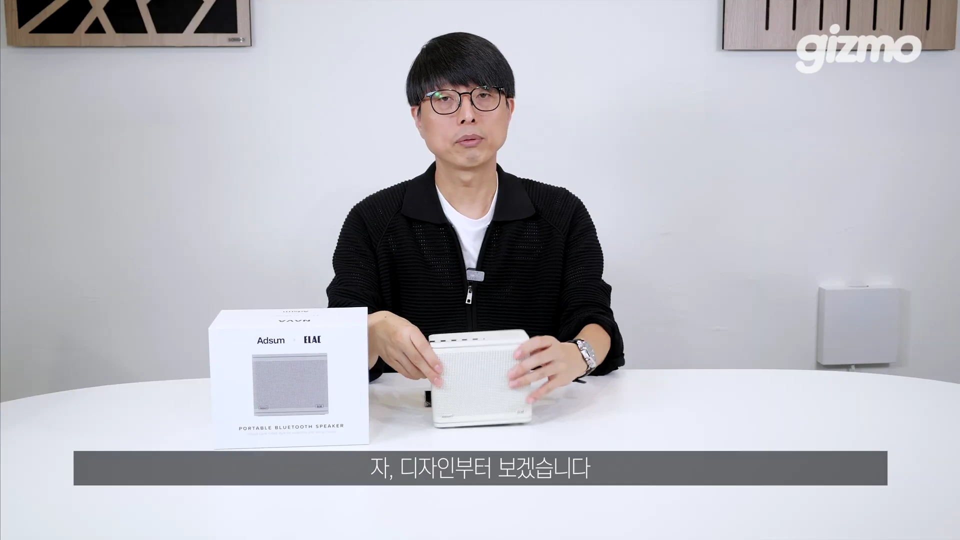 흰색 원형 테이블에 앉은 안경을 쓴 남성이 손으로 작은 크림색 블루투스 스피커를 만지고 있으며, 옆에는 제품 포장 상자가 놓여 있는 스튜디오 영상 장면입니다.