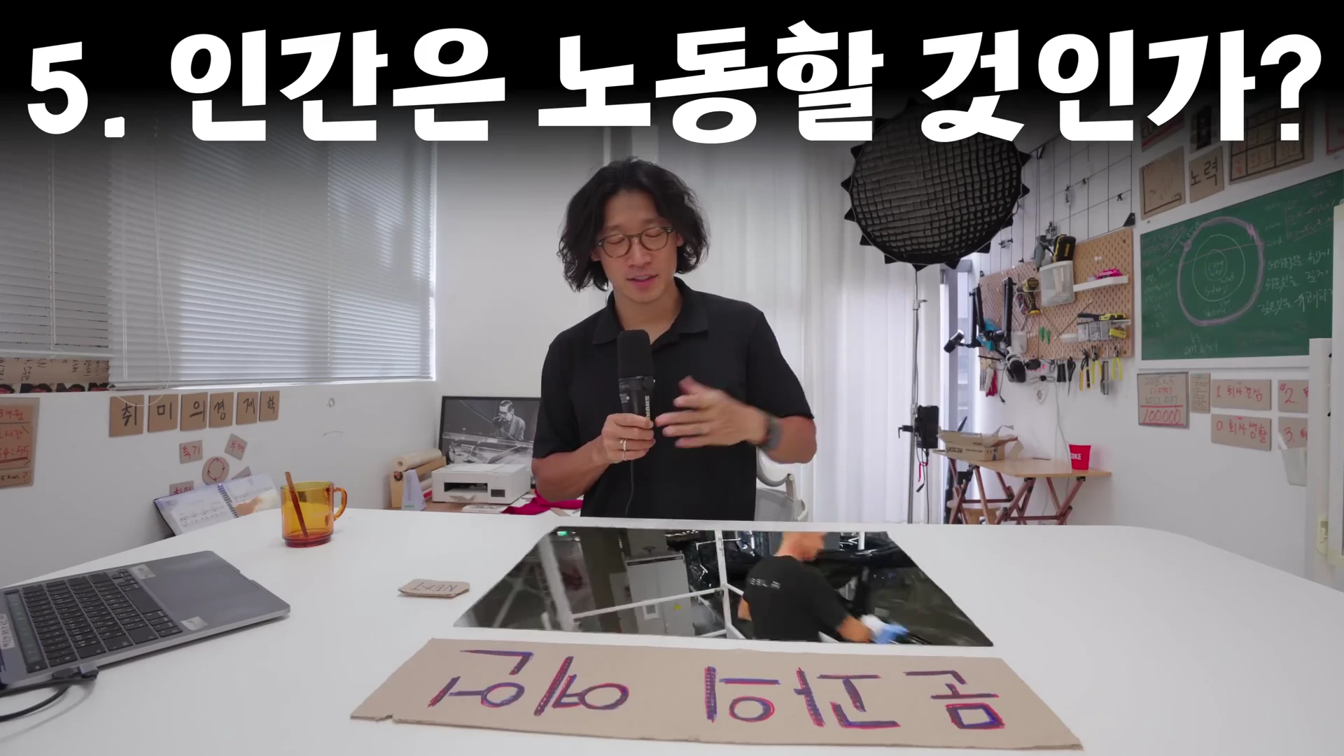 흰색 책상 앞에 앉은 안경 쓴 남성이 마이크를 들고 이야기하고 있으며, 화면 상단에는 '5. 인간은 노동할 것인가?'라는 흰색 문구가 크게 적혀 있습니다.