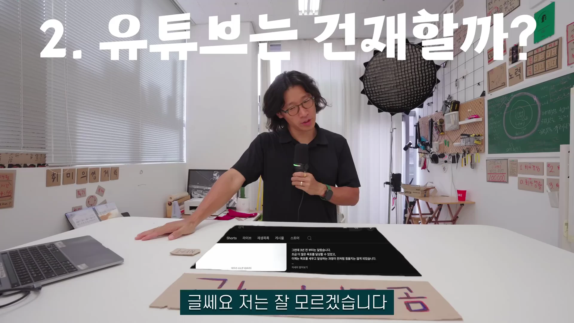 안경을 쓴 남성이 마이크를 들고 작업실 책상 앞에 서서 이야기하고 있으며 화면 상단에는 '2. 유튜브는 건재할까?'라는 자막이, 하단에는 '글쎄요 저는 잘 모르겠습니다'라는 자막이 합성되어 있다.