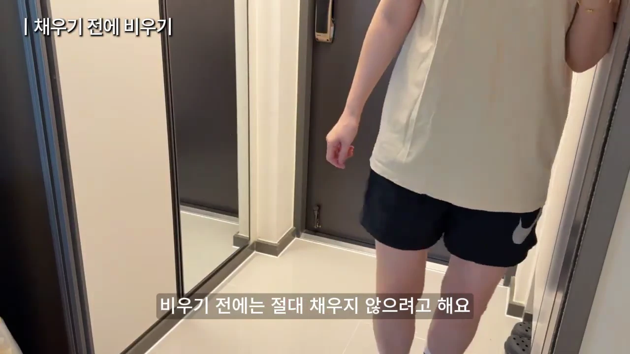 현관에서 베이지색 반팔 티셔츠와 검은색 반바지를 입은 사람이 서 있는 모습