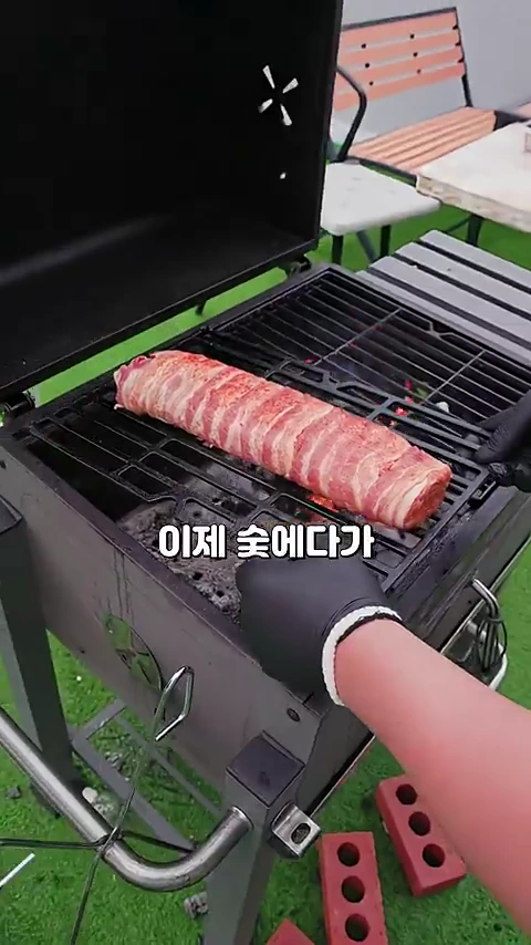 검은색 장갑을 낀 손이 야외 그릴 위에서 베이컨으로 감싼 원통형의 고기를 숯불 쪽으로 옮기고 있는 모습.