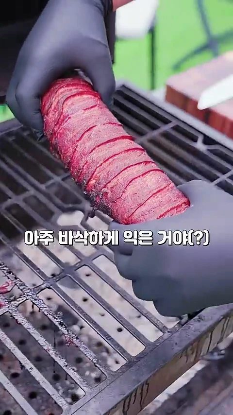 검은색 위생 장갑을 낀 두 손이 숯불 그릴 위에 놓인 길쭉한 고기 롤을 들어 올리고 있는 모습이며, 표면에는 바삭하게 익은 베이컨이 촘촘하게 감겨 있고 중앙에는 '아주 바삭하게 익은 거야(?)'라는 자막이 적혀 있다.