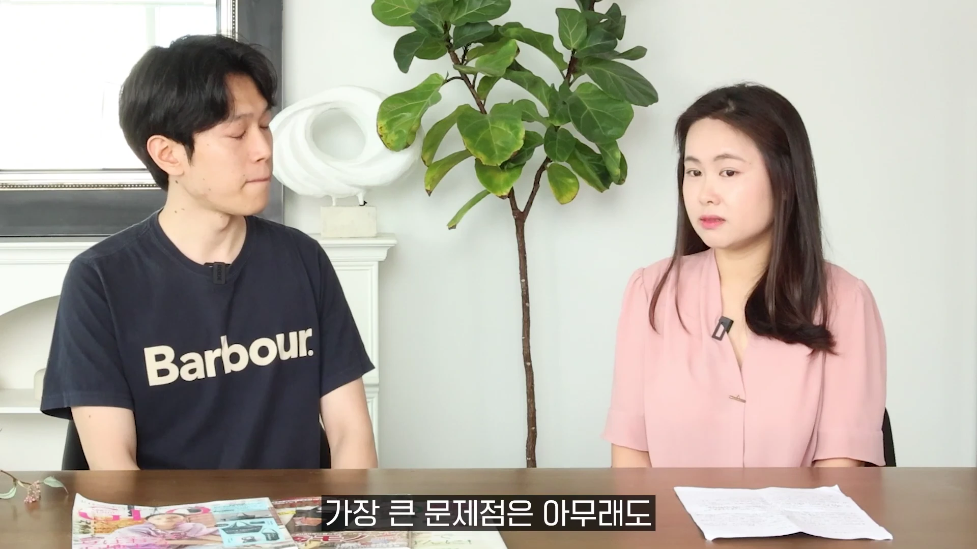 남성과 여성이 테이블에 마주 앉아 진지하게 대화하고 있는 모습으로, 테이블 위에는 잡지와 서류가 놓여 있습니다.